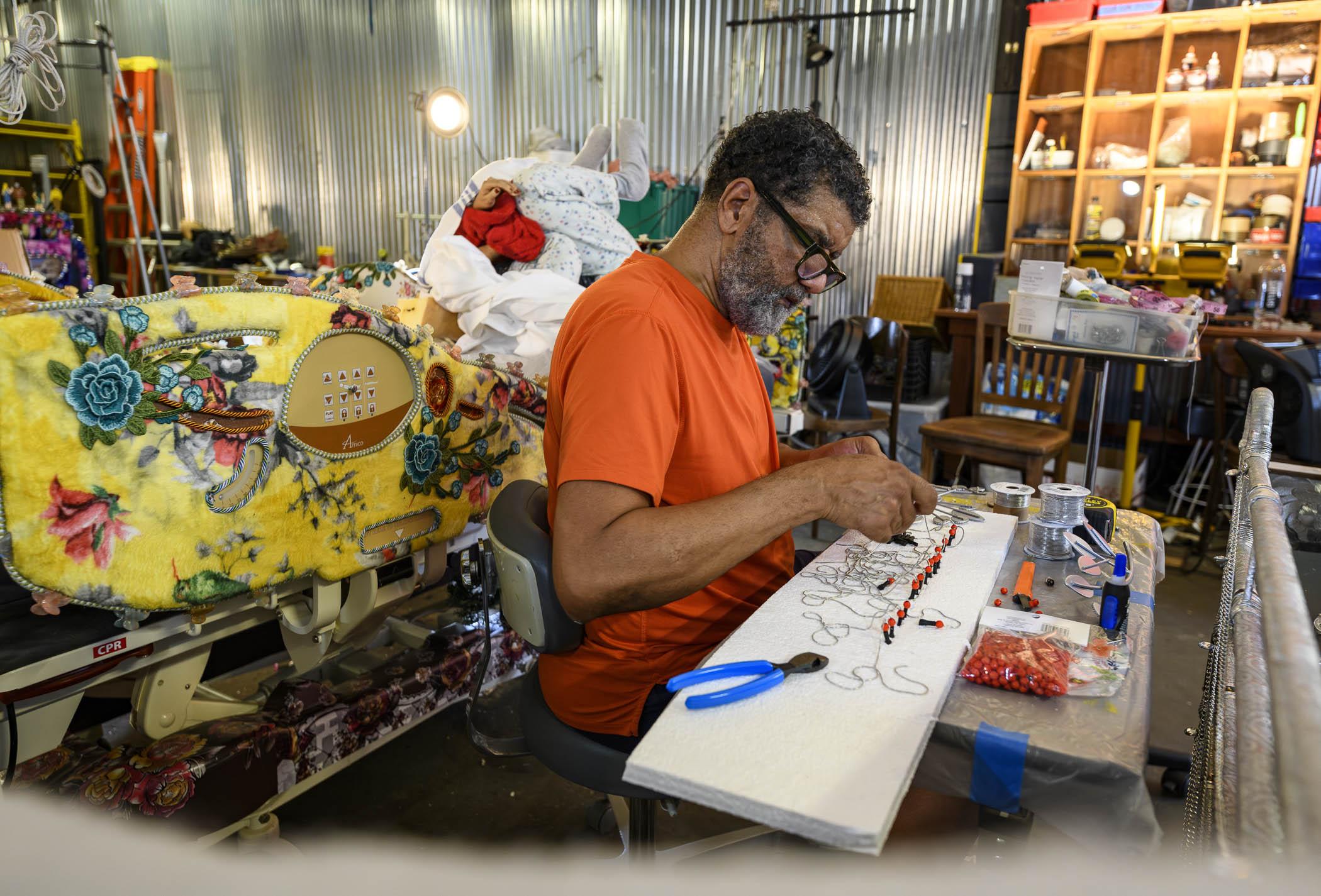 A man wearing glasses and an orange outfit sits at a work table, crafting with wire and beads in a colorful, cluttered studio filled with fabrics and art supplies.
