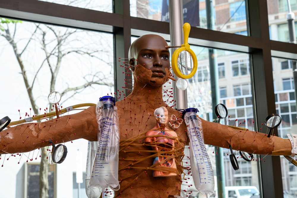 A mannequin with outstretched arms is decorated with red pins, urine collection bags, a toy doll, magnifying glasses, and yellow plastic rings, displayed in front of large windows with urban buildings visible outside.
