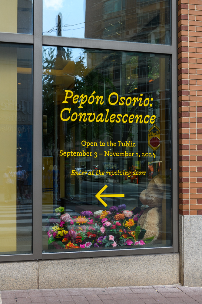A window display advertises Pepón Osorio: Convalescence exhibit, open to the public from September 3 to November 1, 2024. Bright flowers and a large arrow point to the revolving doors for entry.