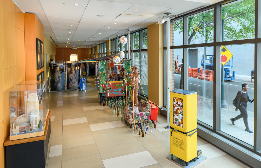 A hallway with large windows on the right displaying street views, and various colorful art installations and sculptures are arranged along the corridor. A person walks outside on the sidewalk.