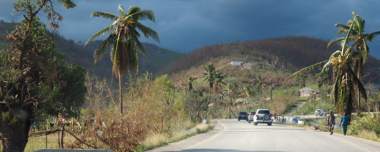 A paved road curves through a rural landscape with palm trees, hills, and scattered houses. Several cars and people are visible under a moody, cloudy sky.