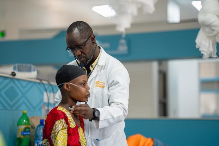 A doctor in a white coat examines a patient wearing a patterned dress and nasal cannula in a medical facility. The doctor uses a stethoscope on the patients back. The setting is modern and brightly lit.