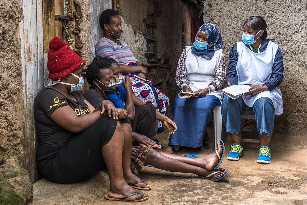 Five women wearing face masks sit together outside a rustic building. Some are seated on the ground, others on chairs, and they appear to be engaged in conversation, with two women holding notebooks or papers.